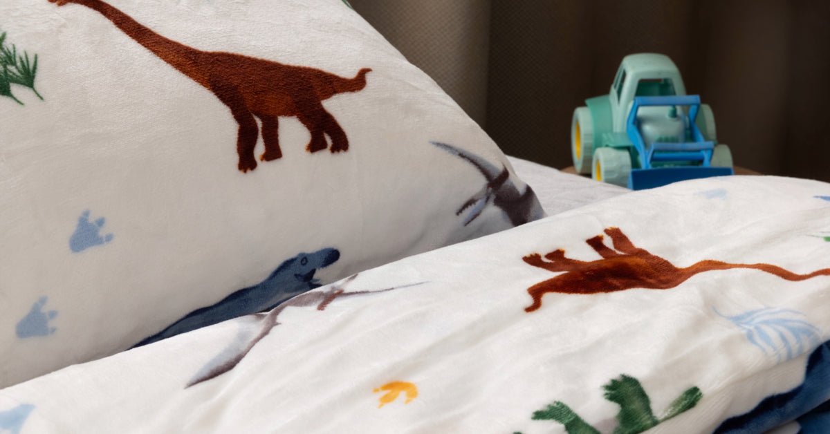 Een close-up van een prachtig opgemaakt bed met een zacht, velvet dekbedovertrek met een dino-thema. De kamer heeft een gezellige uitstraling, met warme kleuren en een houten tafel waarop een schattig autootje staat. De sfeervolle inrichting benadrukt de knusse ambiance van de ruimte. 
foto: dekbedovertrek dino