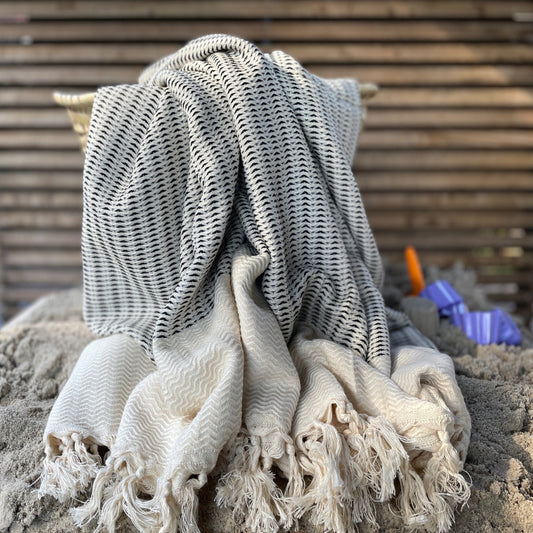 Een hamamdoek zwart ligt op een zandige ondergrond, met schepjes en bakjes op de achtergrond. De doek heeft een verfijnd ontwerp en is perfect voor gebruik in de sauna of op het strand. De schepjes en bakjes, die gedeeltelijk uit het zicht zijn, geven een speelse sfeer aan de afbeelding.
foto: hamamdoek met zand